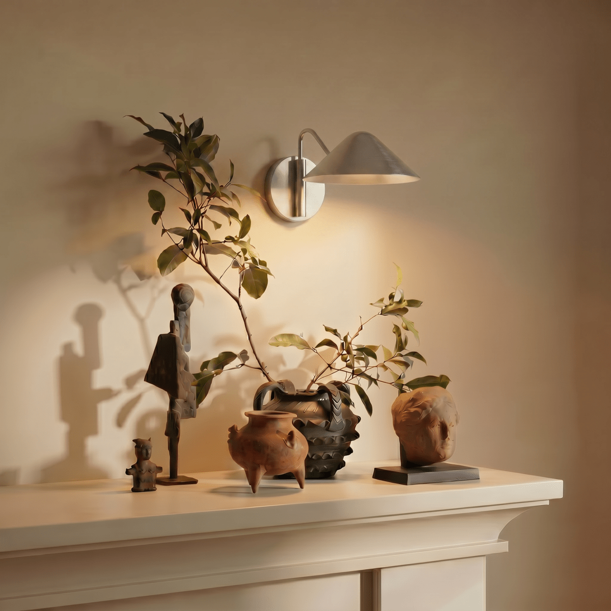 Clérachic chrome cone-shade wall sconce lit above a mantel displaying carved wooden figures and pottery.