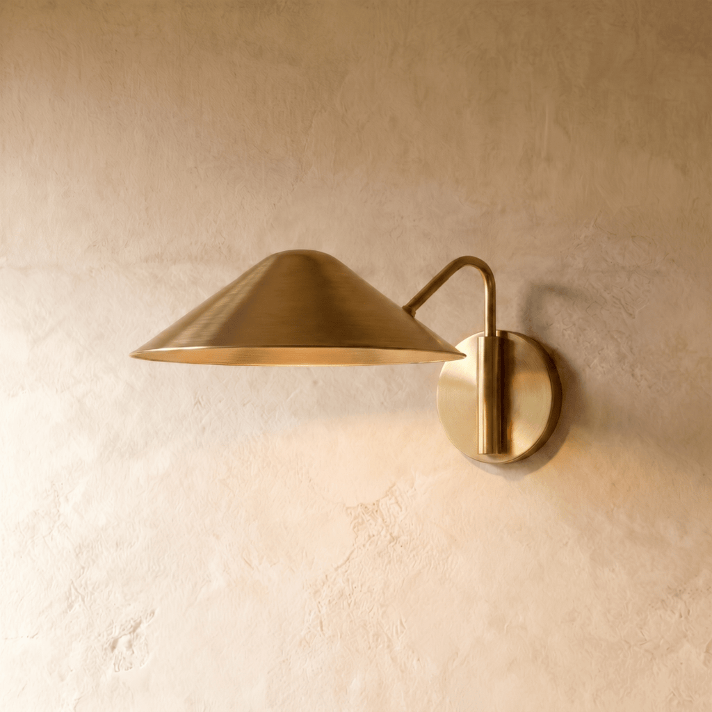 Clérachic cone-shade wall sconce in polished brass with a curved arm, mounted on a plaster wall in studio light.