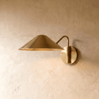 Clérachic cone-shade wall sconce in polished brass with a curved arm, mounted on a plaster wall in studio light.