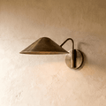Clérachic cone-shade wall sconce in vintage brass with a curved arm, mounted on a plaster wall in studio light.