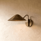 Clérachic cone-shade wall sconce in vintage brass with a curved arm, mounted on a plaster wall in studio light.