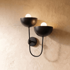 Clérachic wall lamp in matte black, twin dome shades on a looping arm glowing against plaster.