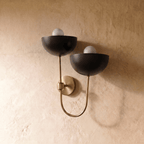 Clérachic wall lamp in brushed brass viewed close, two exposed bulbs glowing under black dome shades.