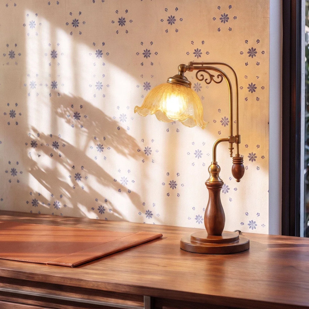 Clérachic table lamp in amber glass glowing on a wood desk beside a leather desk pad and patterned wallpaper.