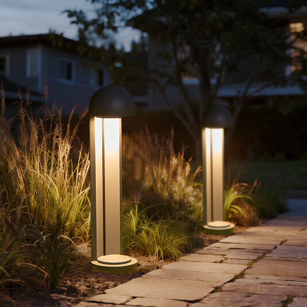 Two bollards lit at dusk among ornamental grasses beside a stone-paved garden path.