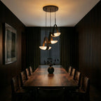 Clérachic three-shade pendant cluster lit above a long dark wood dining table lined with leather chairs in a panelled room.