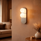 A Clérachic wall light glows warmly beside a sofa nook, its twin globes lighting a vase of dried grass.