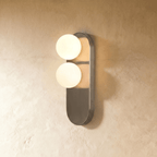 This Clérachic wall light pairs two glowing glass orbs on a brushed steel pill-shaped plate, lit warmly.