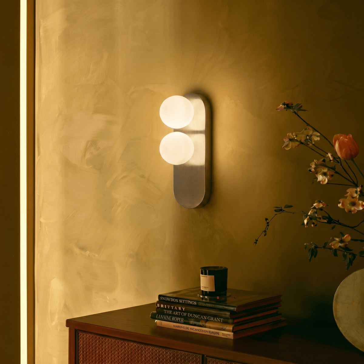 A Clérachic wall light glows warmly above a stack of books and a candle on a dark wood cabinet.