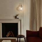 Clérachic wall sconce lit on a pale wall above a fireplace mantel, with a small vase and dried sprig on the shelf below and a floor-length neutral curtain beyond.