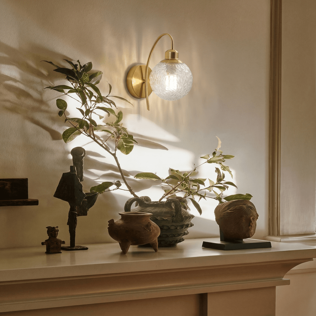 Clérachic wall sconce lit above a pale mantel shelf styled with dark figurines, terracotta-toned pots and a leafy branch casting shadows on the wall.