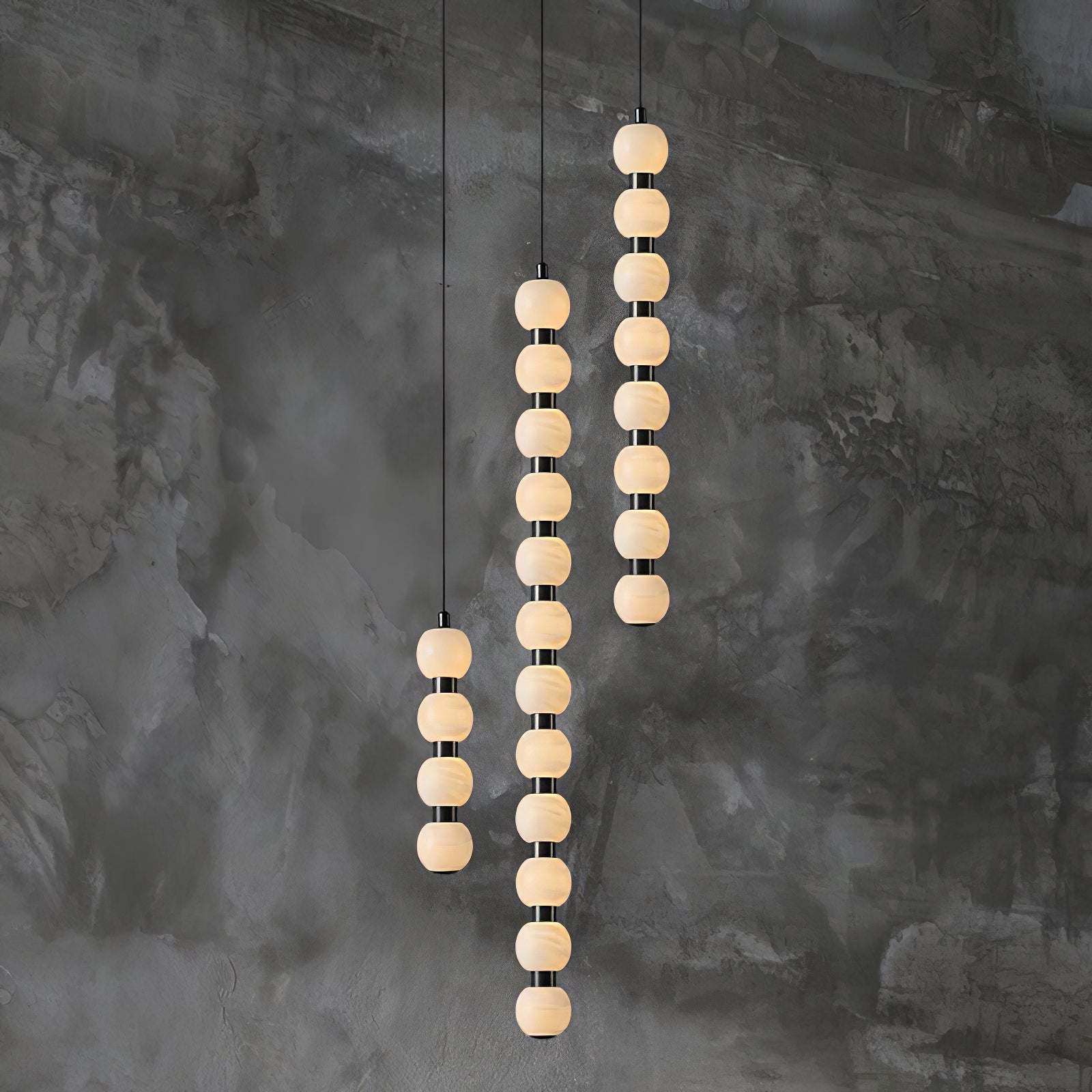 Orbique pendants in three lengths glowing side by side against a dark textured concrete wall