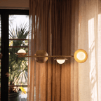 Clérachic brass pendant light glowing above a round dining table with green velvet chairs beside tall windows.