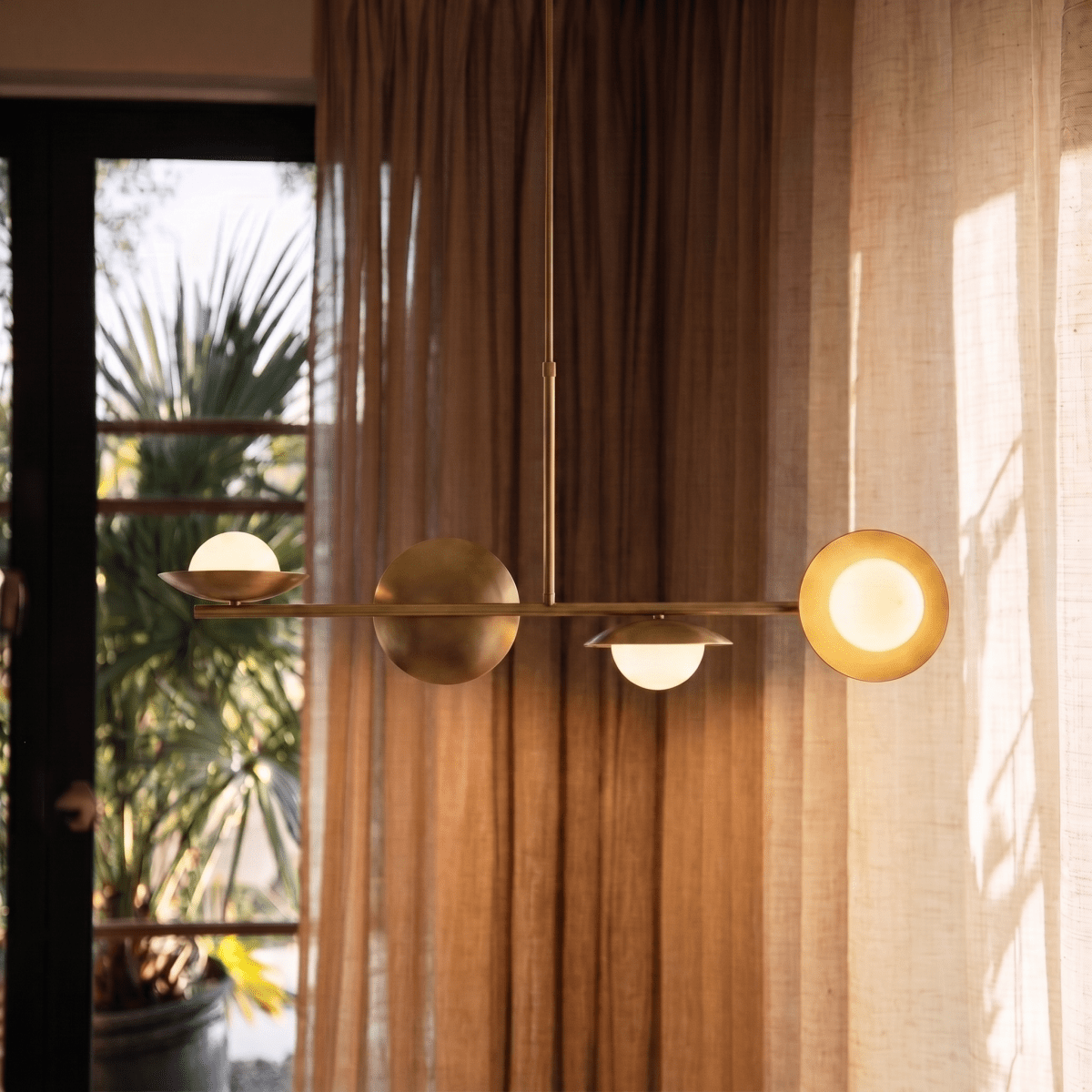 Clérachic brass pendant light glowing above a round dining table with green velvet chairs beside tall windows.