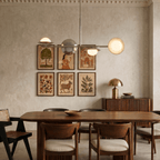 Clérachic chrome pendant light suspended above a dining table, beside a wall of framed folk-art prints in a plaster room.