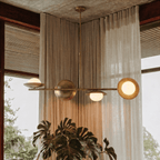 Clérachic vintage brass pendant light glowing above sheer curtains and a large monstera plant near a corner window.