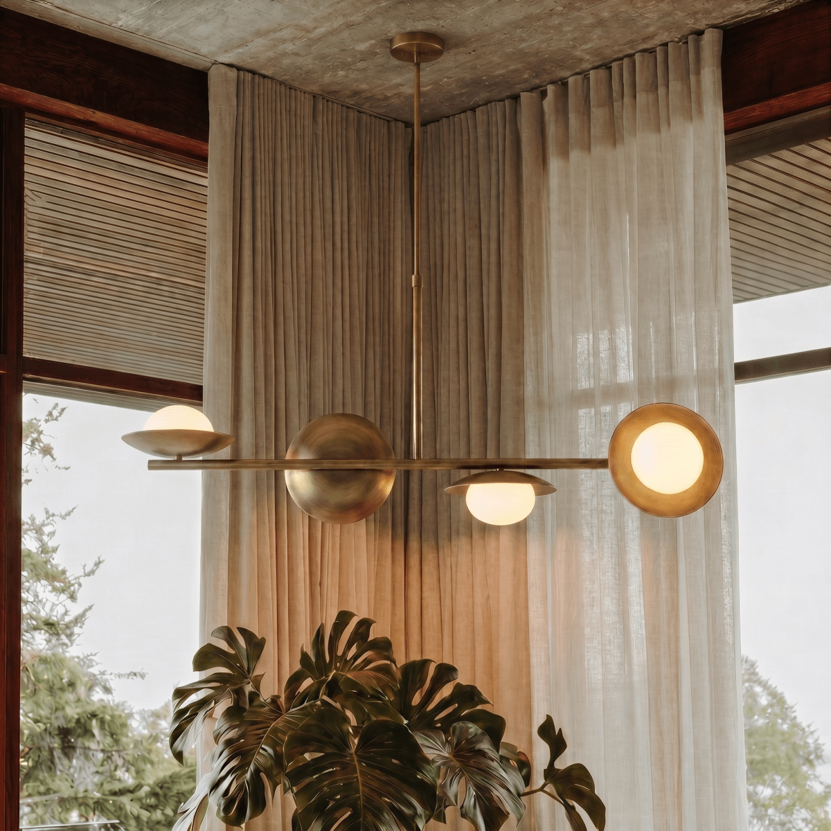 Clérachic vintage brass pendant light glowing above sheer curtains and a large monstera plant near a corner window.