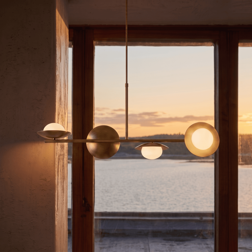 Clérachic vintage brass pendant light silhouetted against a sunset view over water through a timber-framed window.