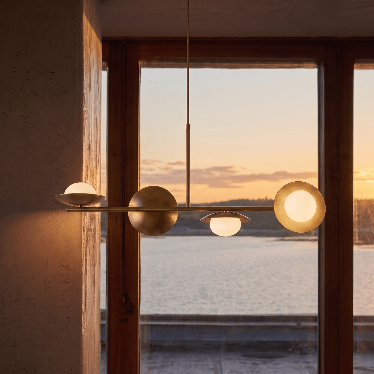 Clérachic vintage brass pendant light silhouetted against a sunset view over water through a timber-framed window.