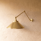 This polished brass Clérachic wall lamp is shown from a different angle, the cone shade glowing warmly.