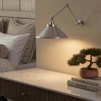 This chrome Clérachic wall lamp lights a marble nightstand holding a bonsai and stacked books.