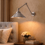 A chrome Clérachic wall lamp lights a tufted headboard beside a nightstand holding roses and books.