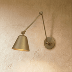 A Clérachic swing-arm wall light in polished brass catches the light on its cone shade against a plaster wall.