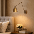 This Clérachic wall light in aged vintage brass lights a tufted headboard bedside table with roses and books.