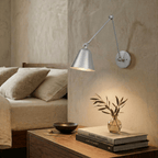 A Clérachic swing-arm wall light in brushed chrome glows beside a bed, lighting a nightstand with books and a vase.