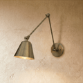 A Clérachic swing-arm wall light in aged vintage brass angles its cone shade down against a bare plaster wall.