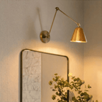 This Clérachic wall light in polished brass hangs above a framed mirror in a marble-tiled bathroom, glowing warmly.