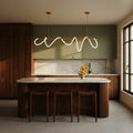 Clérachic Linea chandelier in gold, lit above a marble-topped kitchen island with woven leather stools and sage green cabinetry.