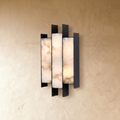 Clérachic marble wall sconce glowing warm amber through three stone columns, mounted on a sand-coloured plaster wall.