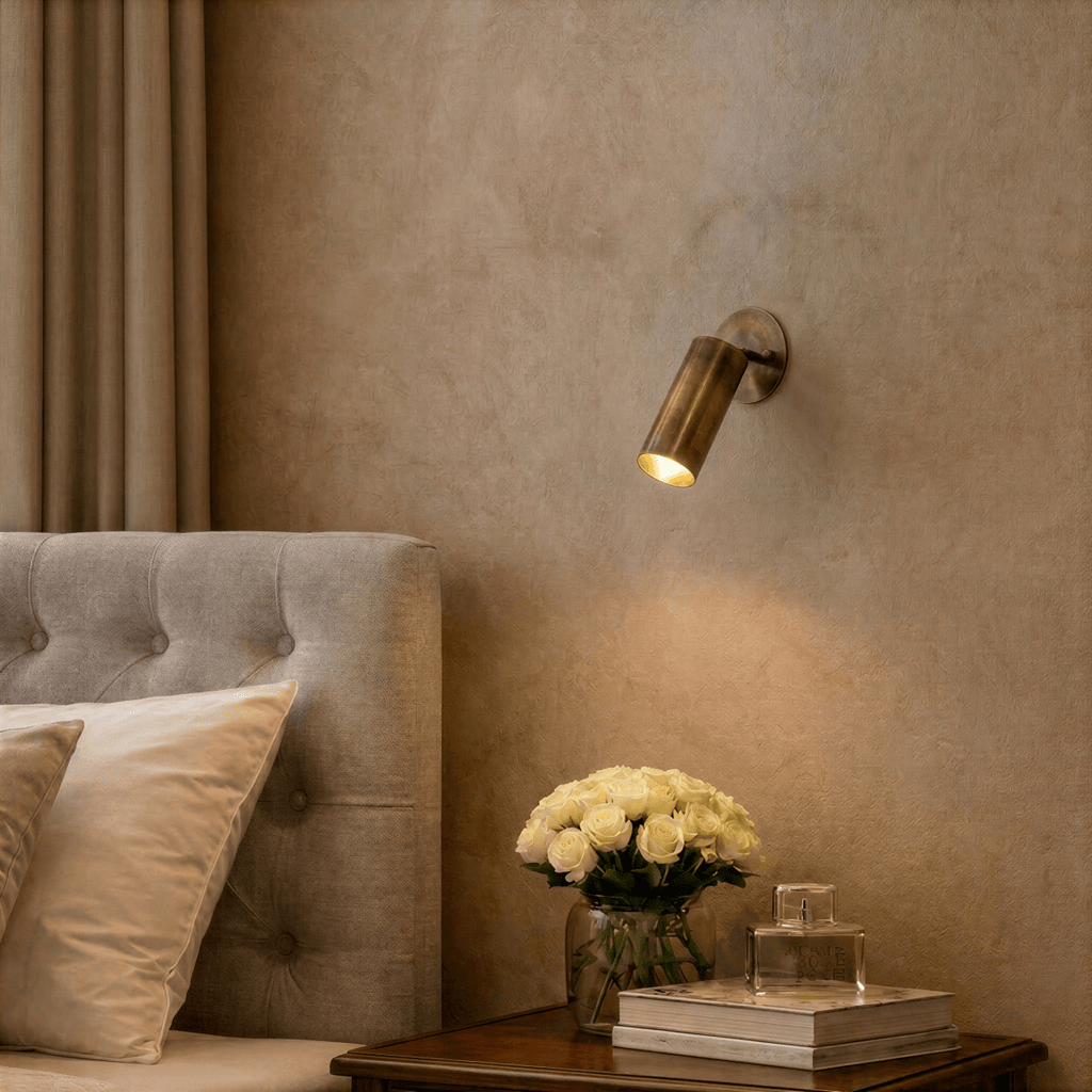 Clérachic wall lamp in brass lighting a bedside table set with white roses and a glass bottle.