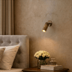 Clérachic wall lamp in brass lighting a bedside table set with white roses and a glass bottle.