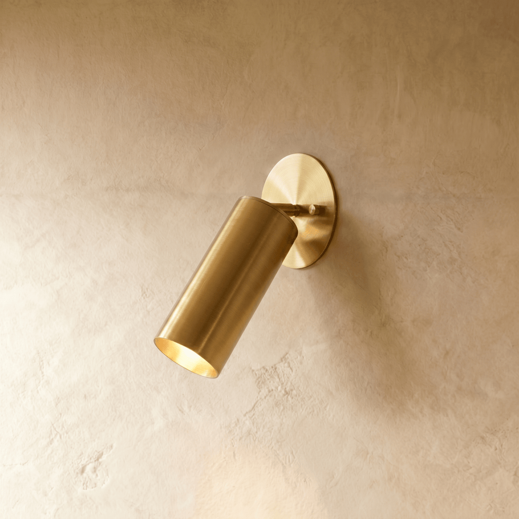Clérachic wall lamp in a warm satin brass finish, the cylinder spotlight angled down from its base.