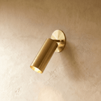 Clérachic wall lamp in a warm satin brass finish, the cylinder spotlight angled down from its base.