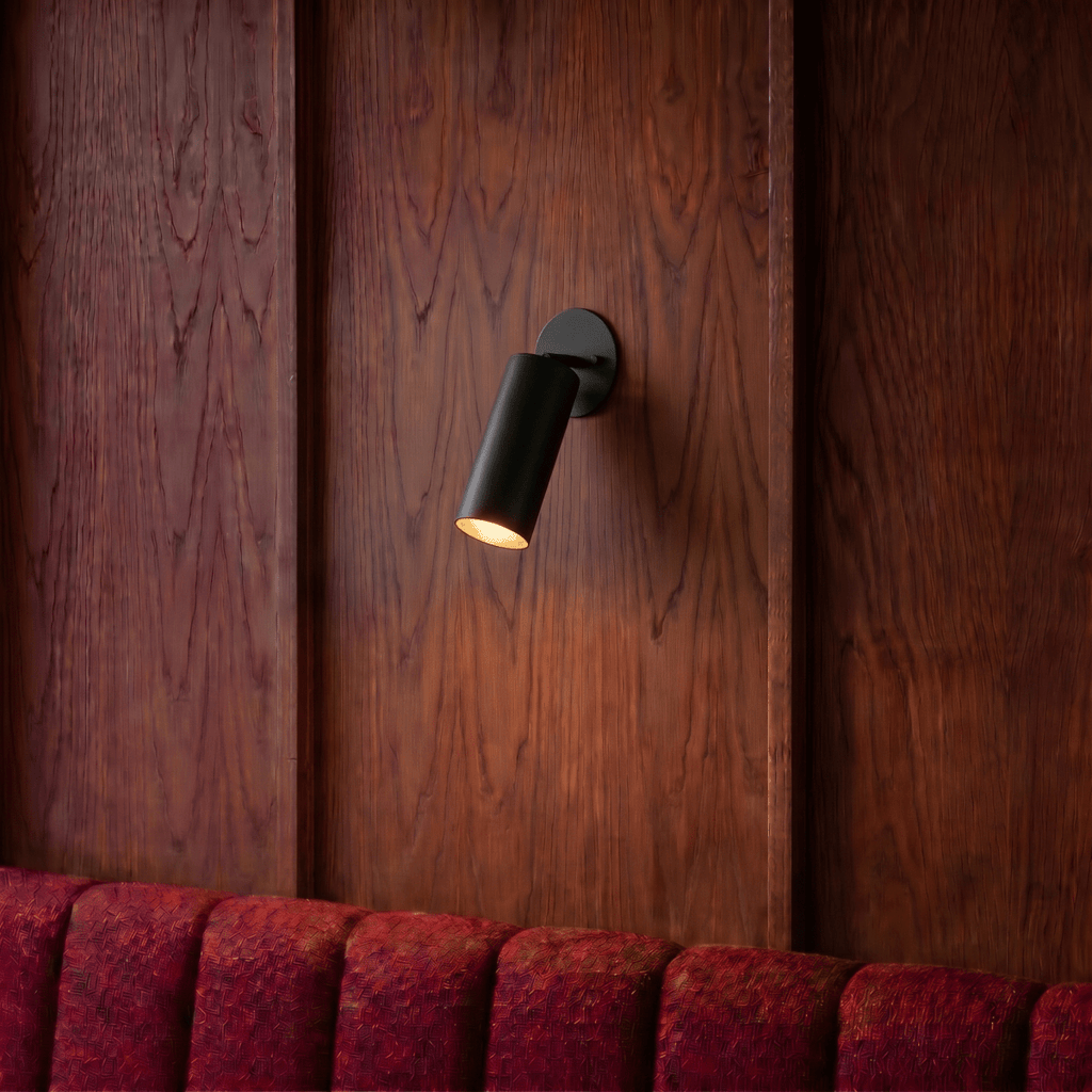 Clérachic wall lamp in matte black mounted on a wood-paneled wall above a red channel-tufted banquette.