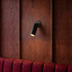 Clérachic wall lamp in matte black mounted on a wood-paneled wall above a red channel-tufted banquette.