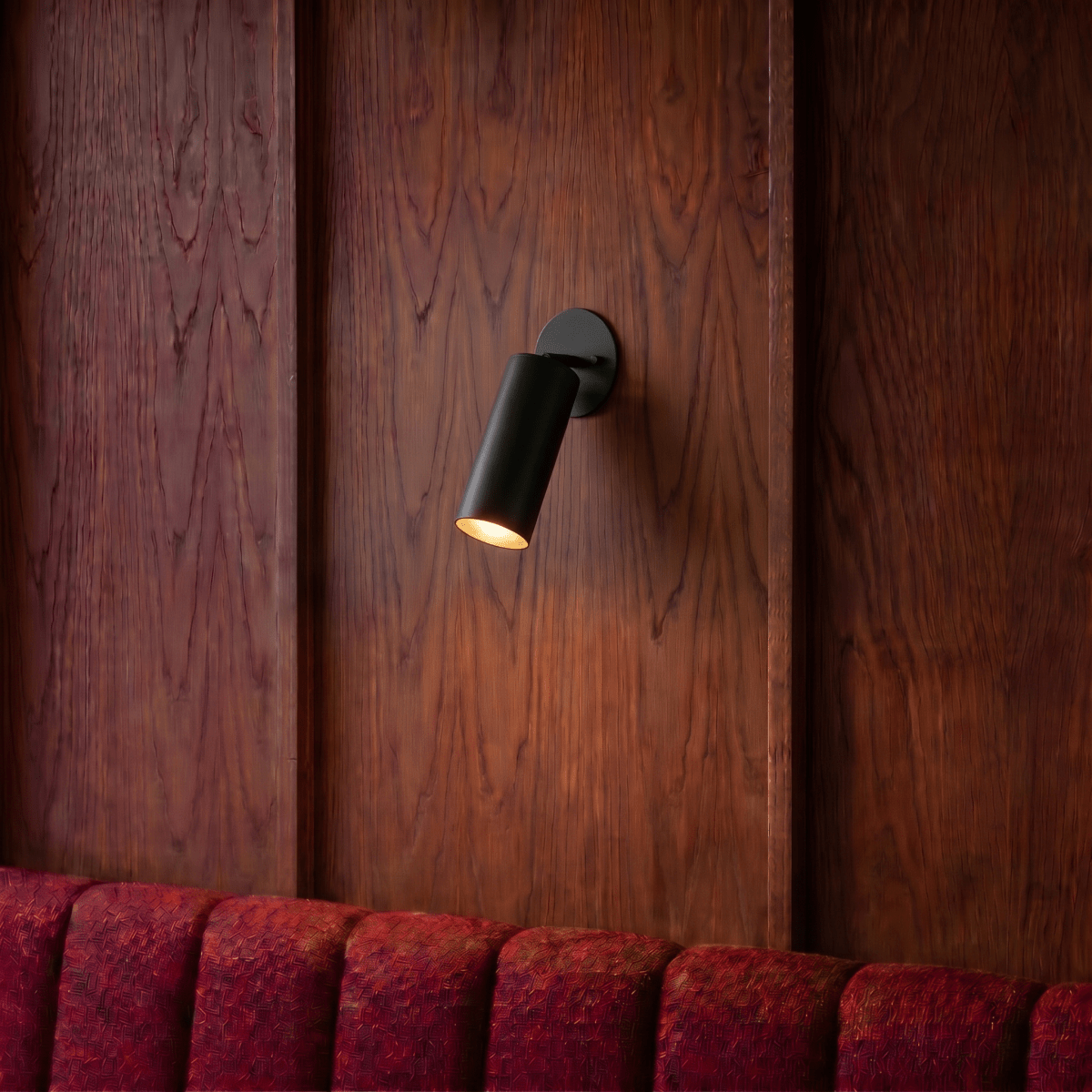 Clérachic wall lamp in matte black mounted on a wood-paneled wall above a red channel-tufted banquette.
