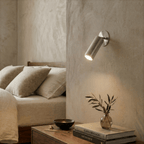 Clérachic wall lamp in chrome lighting a bedside table stacked with books and a small glass vase.