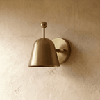A polished brass Clérachic wall lamp viewed from a different angle, the bell shade glowing warmly.