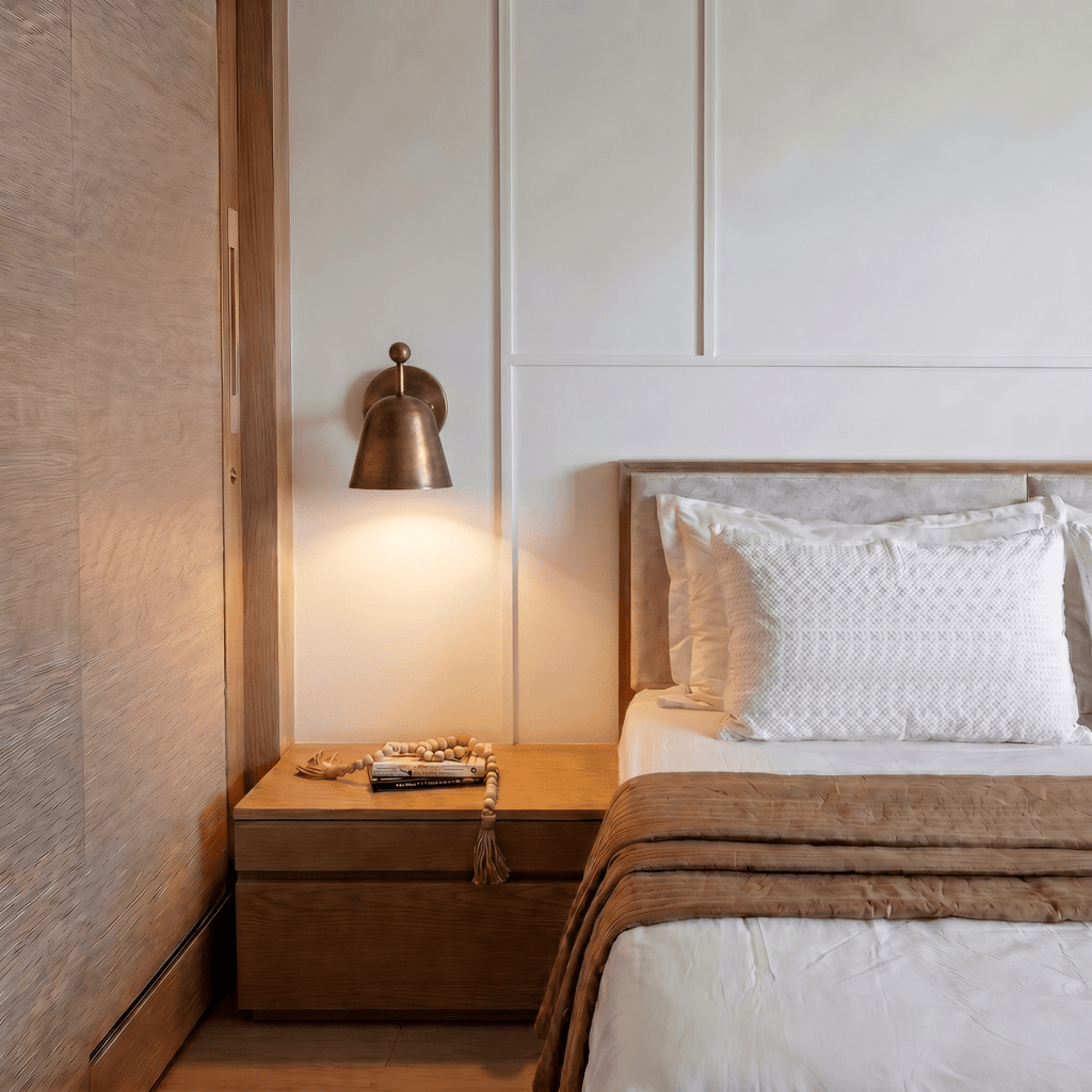 A vintage brass Clérachic wall lamp lights a bedroom nightstand holding a book and prayer beads.