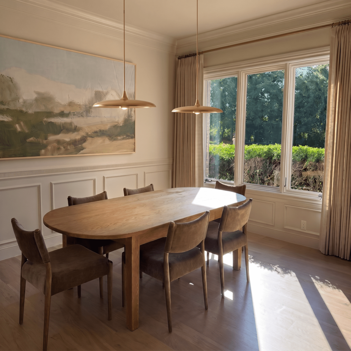 Clérachic brass saucer pendant lights paired above a long oak dining table beneath a large landscape painting.