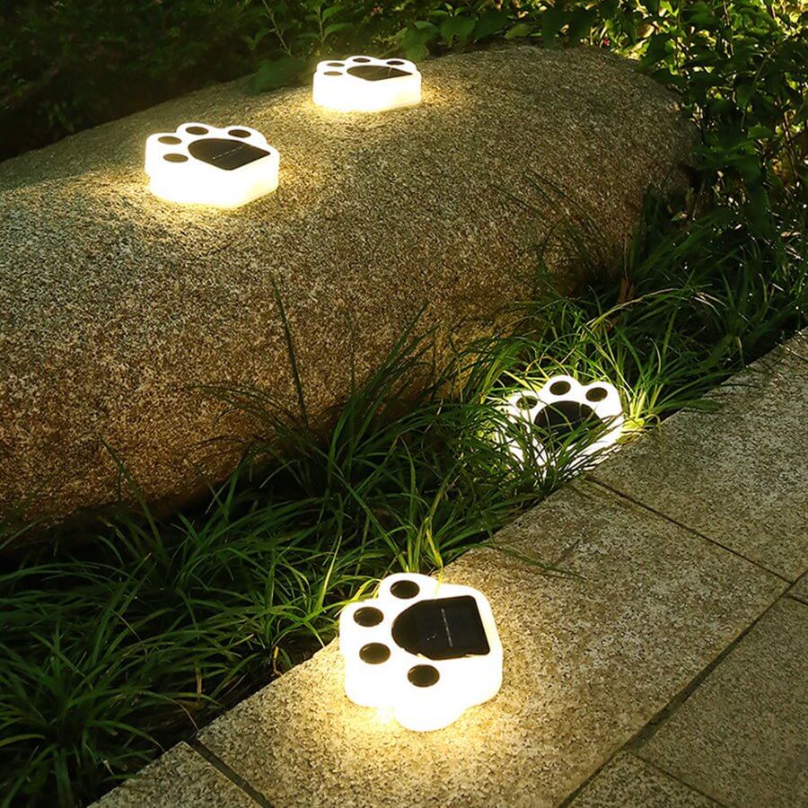 Four paw lights lit warm on a stone boulder and the paving edge beside a border of grass.