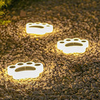 Three paw lights lit warm, resting on a gravel bed with planting at the edge of the frame.