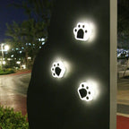 Three paw-shaped step lights glowing white on a dark garden wall at night, with a lit park path beyond.
