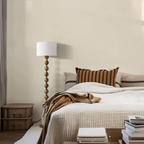 Clérachic stacked-sphere wood floor lamp in walnut, unlit in a daylit bedroom beside a bed dressed in cream quilting and a striped rust cushion.