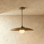 Clérachic dish-shaped pendant light in the Vintage Brass finish, lit, with the opal globe glowing below the shade's rim against a warm plaster wall.