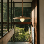 Clérachic dish-shaped pendant light in brass, suspended in a glass-walled sunroom beside a terracotta pot standing on the floor.